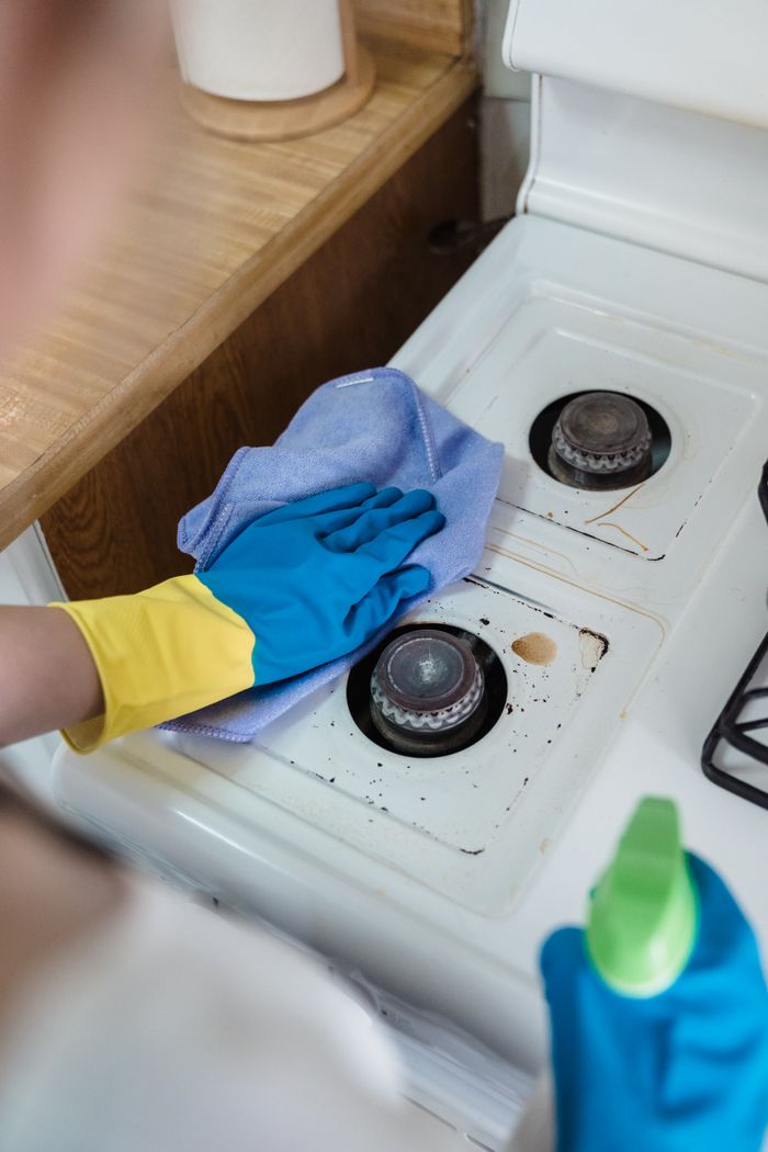services-01 A person wearing rubber gloves cleaning a dirty gas stove with a cloth and spray bottle.
