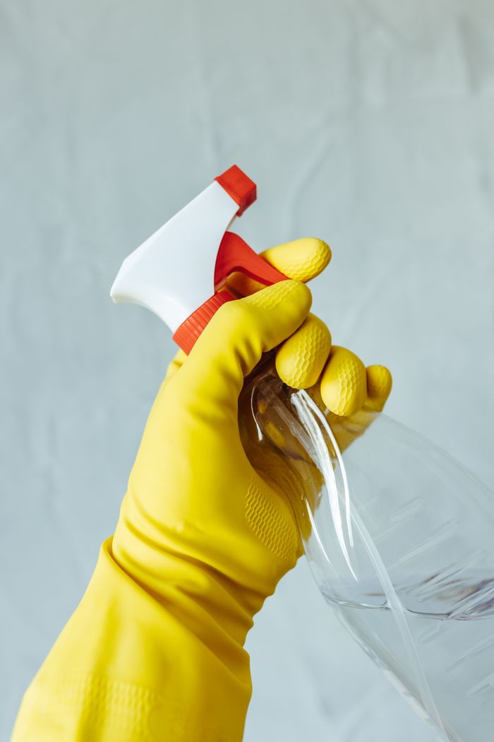 services-02 A hand wearing a yellow rubber glove holds a red and white spray bottle against a neutral background.