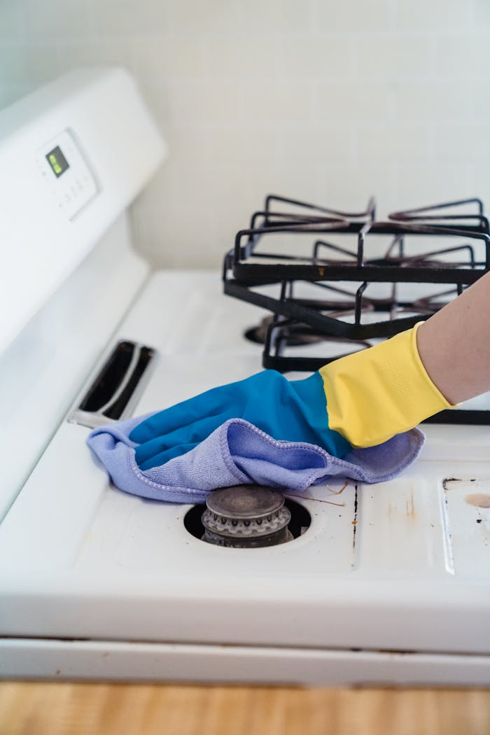 services-03 A close-up of a hand in gloves cleaning a gas stove with a cloth.